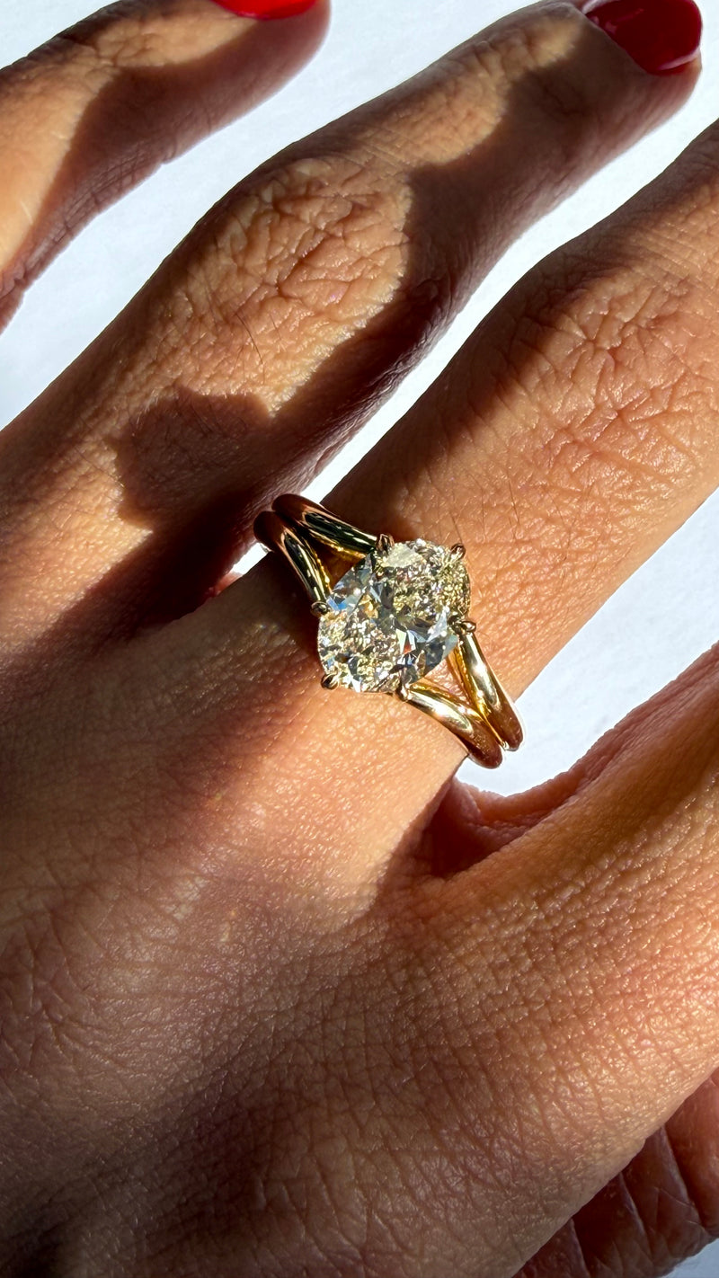A close-up of a hand wearing a yellow gold ring with a large round brilliant-cut diamond. The ring is shown on a finger with red nail polish, set against a bright white background with strong natural shadows.