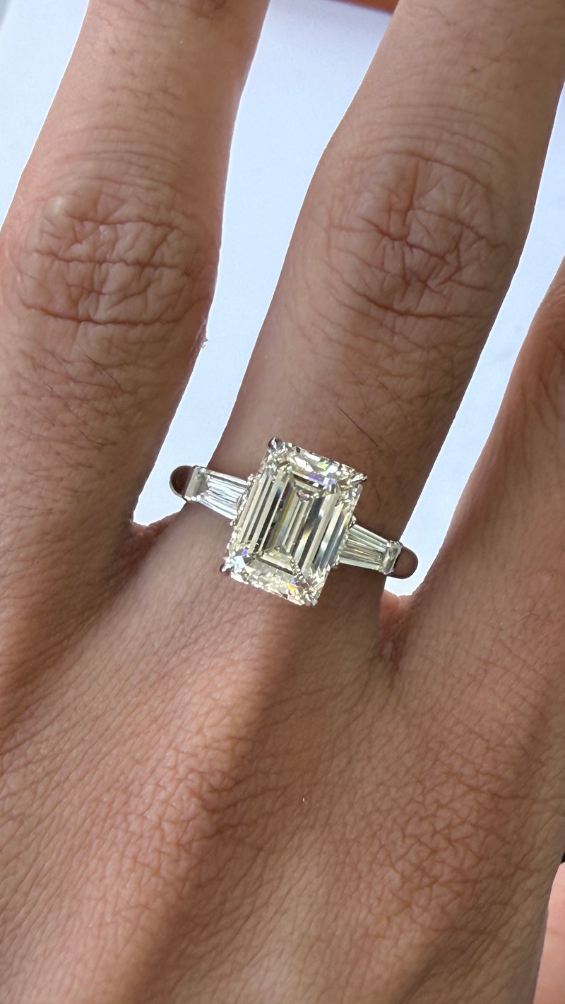An emerald-cut diamond ring with baguette side stones set in a silver-colored band, shown worn on a person's finger.