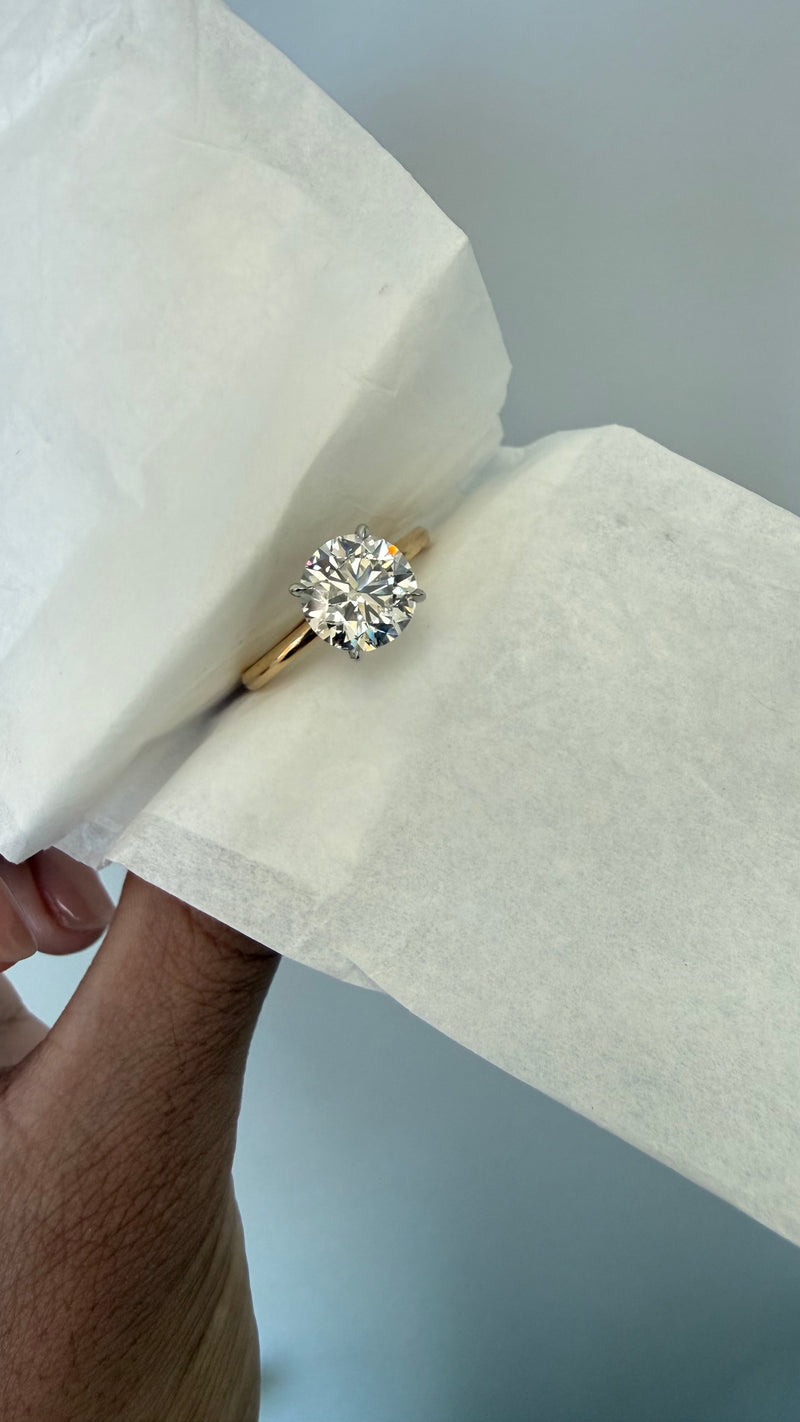 A yellow gold solitaire engagement ring with a round brilliant-cut clear gemstone, held on a piece of white tissue paper by a hand against a light gray background.