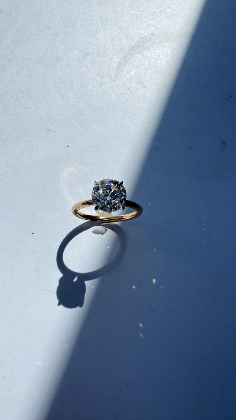 A gold ring with a round-cut clear gemstone, photographed on a blue surface with a strong diagonal shadow.