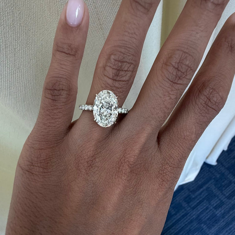 A close-up of a hand with light pink manicured nails wearing a silver-toned oval diamond engagement ring, set against a background of cream fabric and blue carpet.