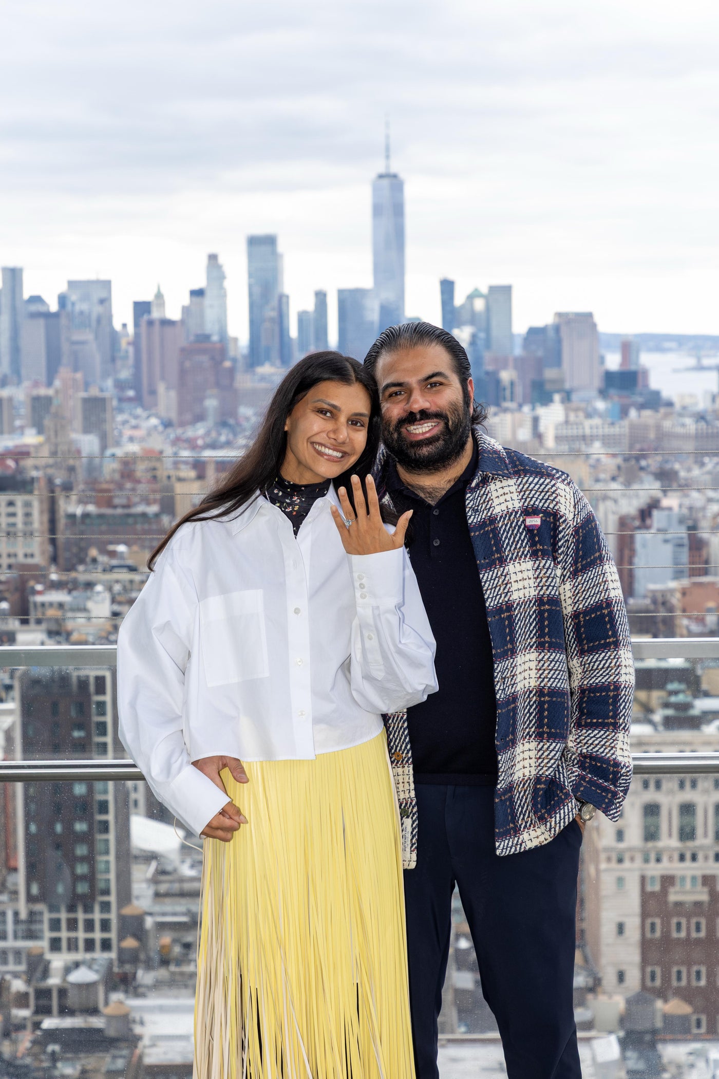 A couple standing on a rooftop with a city skyline in the background holding up her hand with engagement ring
