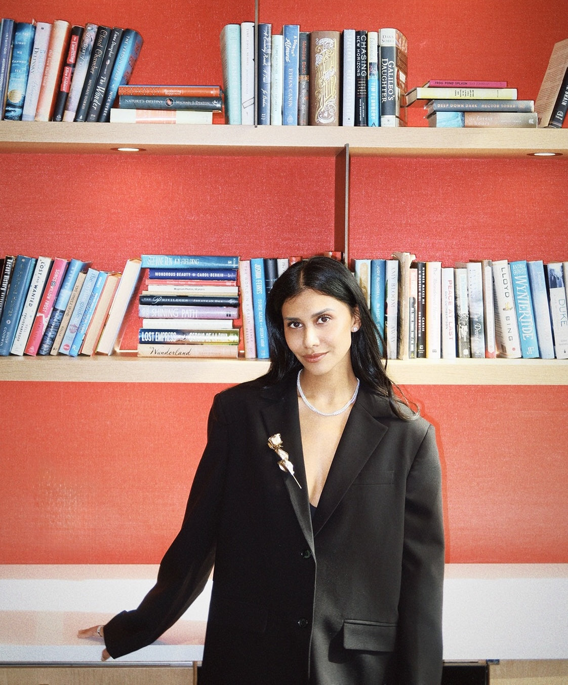 Geeta of van goethem diamonds in a black suit with tennis necklaces standing in front of a bookshelf with red walls.
