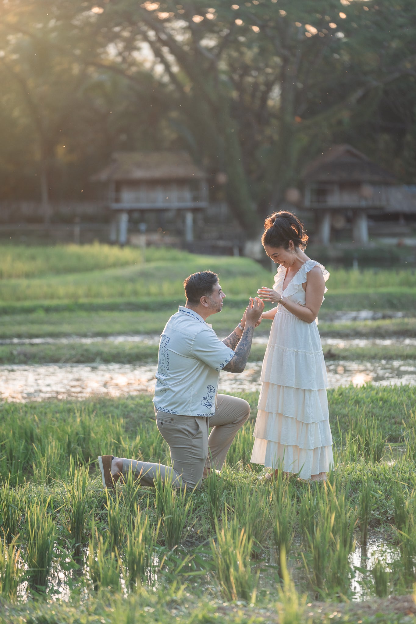 Man proposing to a woman in a scenic outdoor setting with greenery and a building in the background.